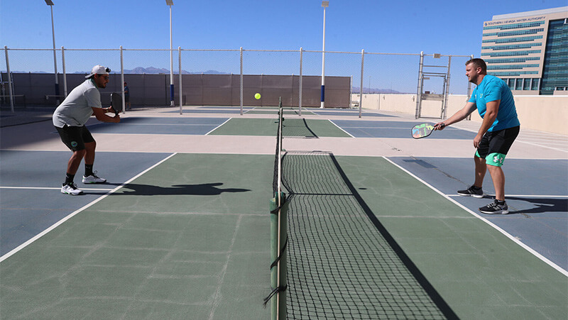 Two men playing pickleball on a court, one serving while the other prepares to return the ball.