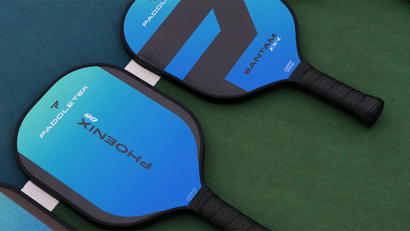 Two blue/black Paddletek Phoenix 66 and Bantam EX-L pickleball paddles place on a green surface